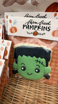 A packaged Frankenstein-themed pumpkin-shaped cookie or sugar cookie with green icing face, black hair, and stitched details, shown in a clear bag with a printed header card that reads 'farm fresh farm fresh PUMPKINS.'.